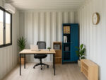 Interior of a neatly organized shipping container office with a minimalist desk, laptop, indoor plants, and vertical storage cabinets under natural light.
